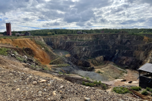 Großer Tagebau in einer Schotterfläche umgeben von Bäumen, Gebäuden und einem Schuppen unter einem bewölkten Himmel, mit verstreuten Steinen und Pflanzen auf dem Boden.