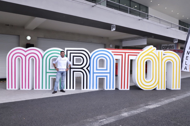Ein Mann steht vor einem großen "Marathon"-Schild mit einem Banner rechts daneben, mit einem Gebäude mit Lichtern, Geländern und Läden im Hintergrund.