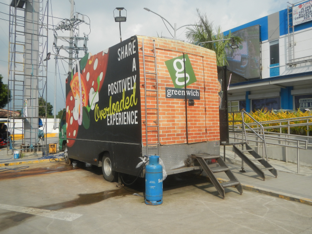 Ein Lastwagen vor einem Gebäude mit einer Tafel, auf der "Teilen Sie eine positiv überladene Erfahrung" steht, Treppen, die hinaufführen, ein Zylinder auf dem Boden, Geländer, Strommasten mit Drähten, Laternen, Bäume und ein bewölkter Himmel im Hintergrund.
