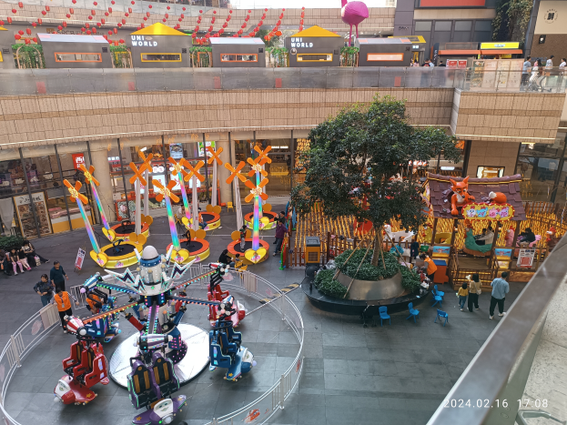 Ein Vergnügungspark innerhalb eines Einkaufszentrums mit verschiedenen Attraktionen, Bäumen, Menschen, Stühlen, Ständen und anderen Gegenständen, mit Gebäuden oben und einem Wasserzeichen unten rechts.