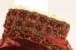 Nahaufnahme eines roten und goldenen Kleides mit aufwändiger Spitzenverzierung auf einem Mannequin vor einem weißen Hintergrund.