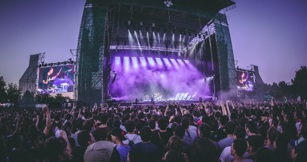 Eine große Menschenmenge versammelt sich auf einem Musikfestival mit einer Bühne, auf der Musiker spielen, beleuchtet von Scheinwerfern und umgeben von Bildschirmen, vor einer Kulisse aus einem Gebäude, Bäumen und einem klaren blauen Himmel.