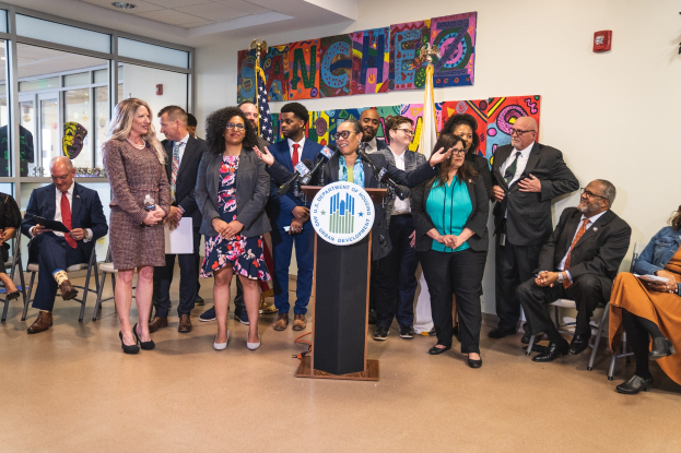 Gruppe von Menschen vor einem Rednerpult mit Mikrofon und einer Tafel mit der Aufschrift "Sancho City Councilman Joins City Council", mit sitzenden und stehenden Teilnehmern, Flaggen, Feuerlöscher und Deckenbeleuchtung im Hintergrund.