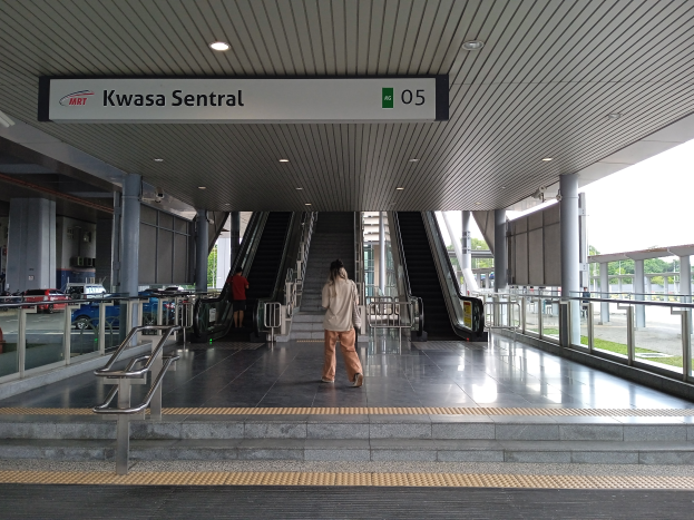 Eine Frau geht eine Rolltreppe in einem Bahnhof hinunter, umgeben von Geländern, Säulen, Texttafeln, Deckenleuchten, Glaswänden, Straßenfahrzeugen, Bäumen und einem klaren blauen Himmel.