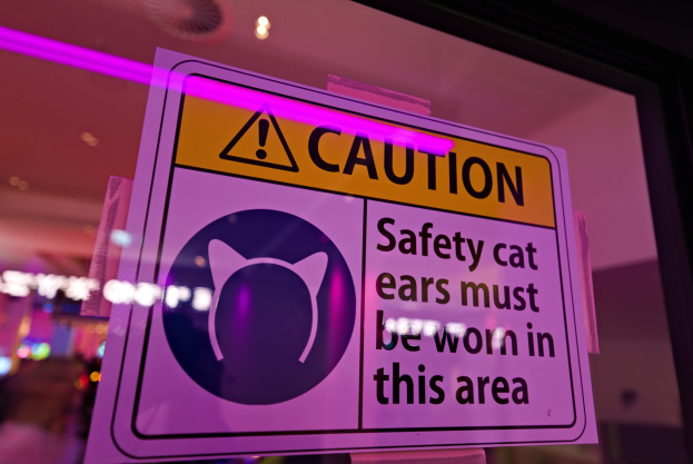 Hellgelbes Warnschild in einem Ladenschaufenster mit der Aufschrift "Vorsicht: Sicherheitskatzenohren müssen in diesem Bereich getragen werden" vor einem unscharfen Hintergrund.