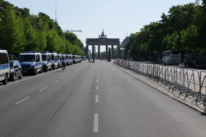 Eine lange Reihe von Polizeiwagen, die auf der Seite einer Straße vor dem Brandenburger Tor in Berlin, Deutschland, geparkt sind, mit Menschen, die Fahrräder fahren und auf der Straße stehen, Absperrungen und Bäume an den Seiten und ein Tor mit Statuen im Hintergrund.