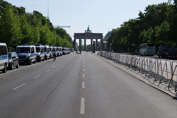 Eine lange Reihe von Polizeiwagen, die auf der Seite einer Straße vor dem Brandenburger Tor in Berlin, Deutschland, geparkt sind, mit Menschen, die Fahrräder fahren und auf der Straße stehen, Absperrungen und Bäume an den Seiten und ein Tor mit Statuen im Hintergrund.