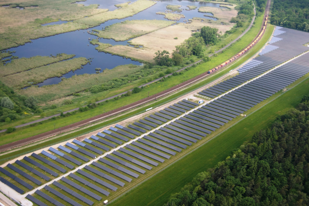 Eine Luftaufnahme eines Solarparks mit Panelen in einem Feld, umgeben von Bäumen, Gras, Pflanzen und Wasser, mit einem Zug auf einer Eisenbahnschiene in der Nähe.