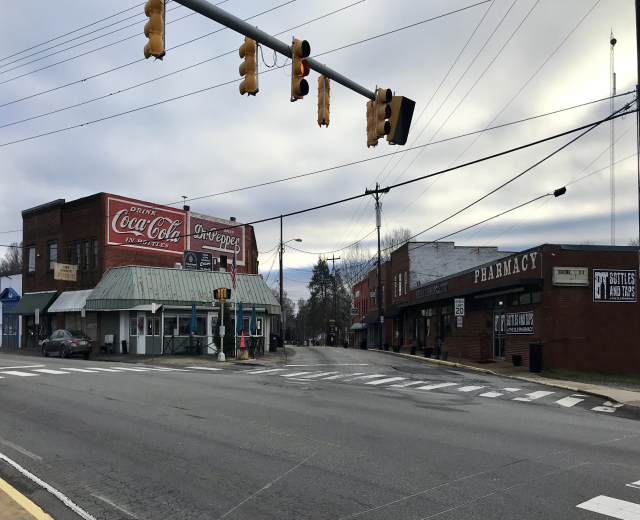 Stadtstraßenkreuzung mit Ampel, Fahrzeugen, Gebäuden mit Schildern, Strommasten mit Drähten, Bäumen und einem bewölkten Himmel.