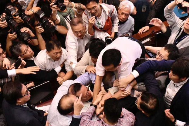 Eine Gruppe von Menschen steht zusammen vor einer Menge, einige halten Kameras, im Hintergrund sind Tische mit Papieren und anderen Gegenständen zu sehen, was auf eine Diskussion im Zusammenhang mit der Hongkonger Börsenschließung aufgrund der Coronavirus-Pandemie hindeutet.