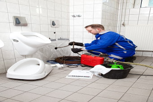 Ein Mann in einer blauen Uniform sitzt vor einem Urinal auf dem Boden und hält eine Pfeife in der Hand, umgeben von Reinigungswerkzeugen und -mitteln, mit Urinalen links und einem Fenster rechts.
