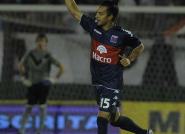 Ein Fußballer auf dem Boden, der ein Tor feiert, mit einer unscharfen Menge und einer weiteren Person im Hintergrund hinter einer Texttafel.