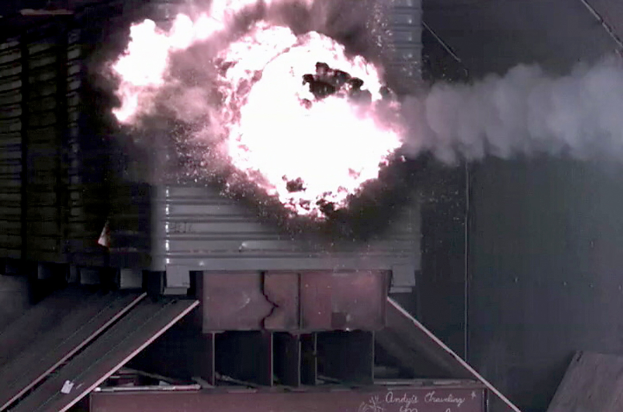 Große Explosion in der Mitte eines Gebäudes mit Rauch, einer Wand im Hintergrund und Metallobjekten mit einer Tafel unten.