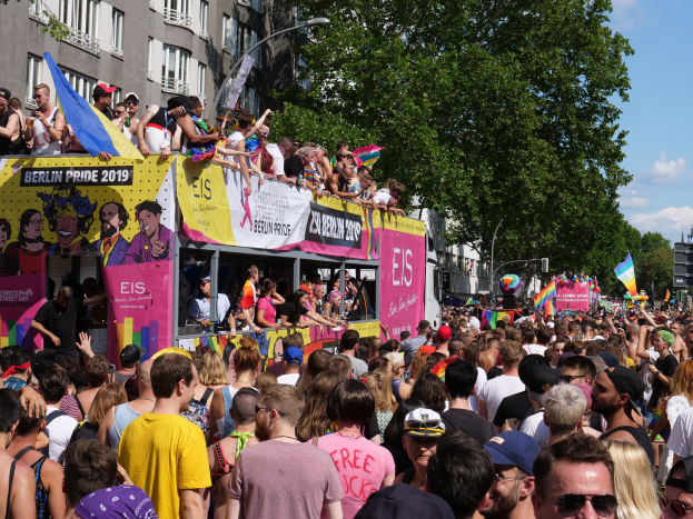 Eine große Menschenmenge, die die Straße bei der Christopher Street Day Parade in Berlin 2019 entlanggeht, viele mit Mützen und Schutzbrillen, Fahnen haltend, mit Bannern und Gebäuden im Hintergrund unter einem bewölkten Himmel.