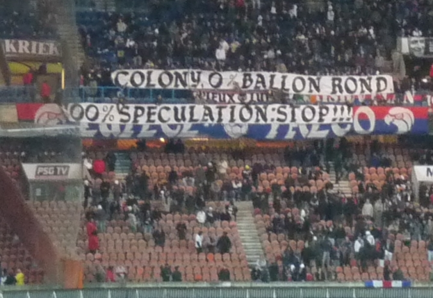 Eine große Menschenmenge in einem Stadion mit Bannern und Stühlen im Vordergrund, einem Zaun unten und Treppen auf der linken Seite.