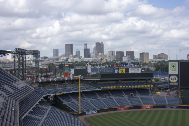 Baseballstadion mit einer Stadtkulisse im Hintergrund, gefüllt mit Stühlen, Pfosten und Brettern, auf Gras gesetzt mit Bäumen und Gebäuden in der Ferne unter einem bewölkten Himmel.