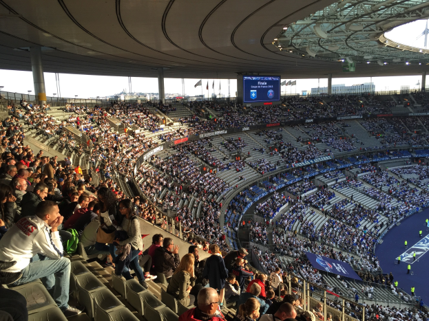 Große Menschenmenge in einem Stadion bei einem Fußballspiel, mit einer Bühne rechts, Fahnen, Masten, einem Bildschirm und der Allianz Arena in München, Deutschland im Hintergrund.