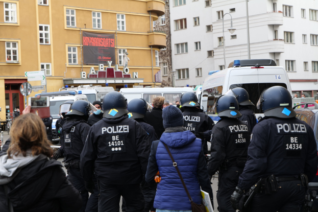 Polizeibeamte in Uniform stehen einer Menge von Helm- und Jackenträgern gegenüber während einer Demonstration in Berlin, Deutschland, mit Fahrzeugen, Gebäuden, Laternenpfählen und einem Banner im Hintergrund, während eine Person eine Kamera hält.