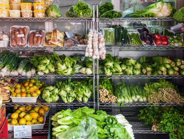 Ein Gang in einem Supermarkt mit frischem Gemüse und Obst, verpackten Artikeln unter Plastik, Kartons, einer Texttafel und einem Glasfenster im Hintergrund.