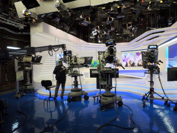 Ein Mann in einer Live-Broadcast-Umgebung hält ein Mikrofon vor einem Fernseher, umgeben von Kameras, einem Stuhl, einem Anzeigebildschirm und Deckenleuchten.
