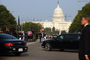 Trauerzug mit Menschen in Mützen und Fahnen vor dem Kapitolsgebäude, Fahrzeuge auf der Straße, Laternenpfähle, Bäume und Gebäude mit Fenstern unter einem klaren Himmel.