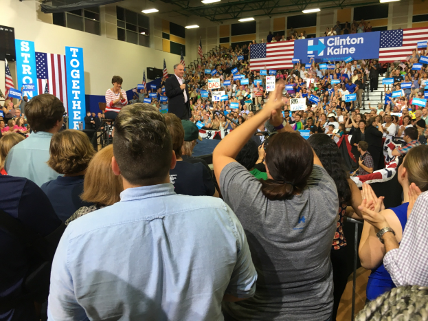 Eine große Menge applaudiert und jubelt vor einer Bühne, auf der ein Mann in ein Mikrofon spricht, während eine Frau neben ihm steht; Fahnen, Banner und ein Lautsprecher sind auf dem Bühnenhintergrund zu sehen.
