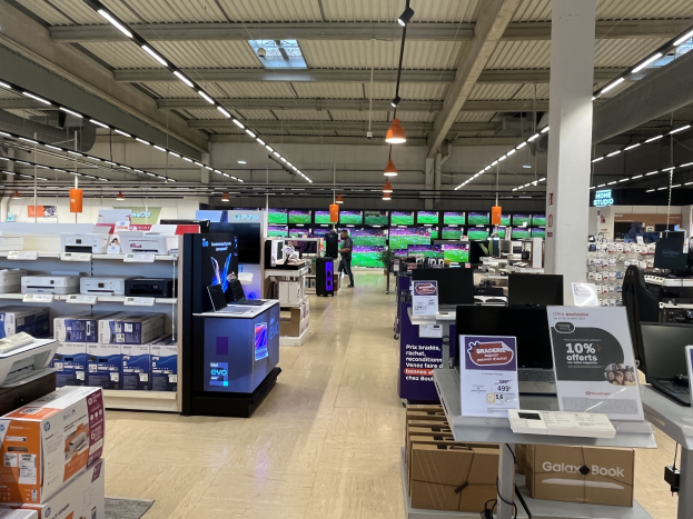 Großer Raum mit Elektronik einschließlich Computern, Druckern und Kisten, die in Regalen angeordnet sind, Menschen auf dem Boden, eine Säule, Schilder, Deckenlampen und Deckenventilatoren in einem Walmart-Laden.