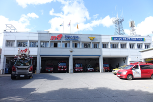 Feuerwache mit mehreren Feuerwehren vor dem Gebäude geparkt, ein Gebäude mit Fenstern und Text, Flaggen auf Stangen im Hintergrund, bewölkter Himmel und eine ferne Turm.