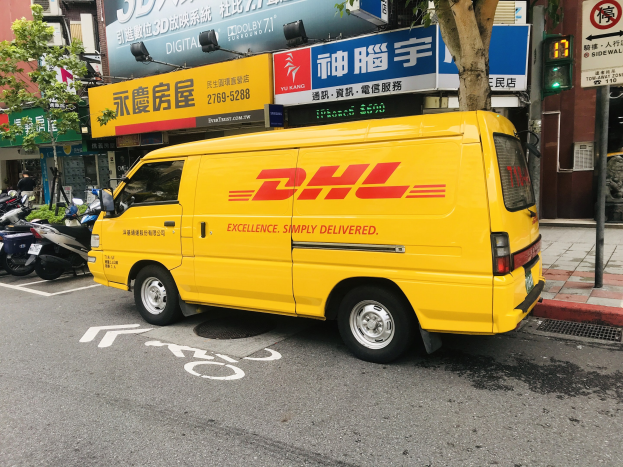 Gelbes DHL-Lieferfahrzeug neben Gebäuden, Bäumen, Schildern, Mästen, Ampeln und einer Person auf dem Gehweg geparkt.
