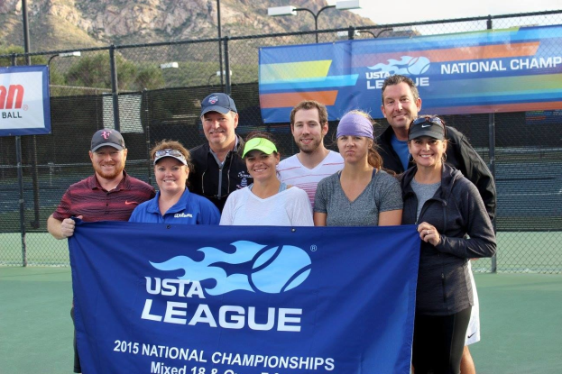 Gruppe von Menschen auf einem Tennisplatz, die eine "2015 USTA National Championships"-Fahne halten, mit Zäunen, Fahnen, Laternenmasten, Bäumen, Bergen und einem klaren blauen Himmel im Hintergrund.