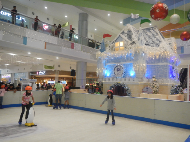 Eine Gruppe von Menschen, die auf einer Eisbahn in einem Einkaufszentrum Schlittschuh laufen, umgeben von einem hausähnlichen Aufbau, der mit Lichtern und festlichen Dekorationen geschmückt ist, mit Menschen in der Nähe, Geländern, Säulen, Texttafeln und Deckenleuchten, die eine festliche Atmosphäre schaffen.