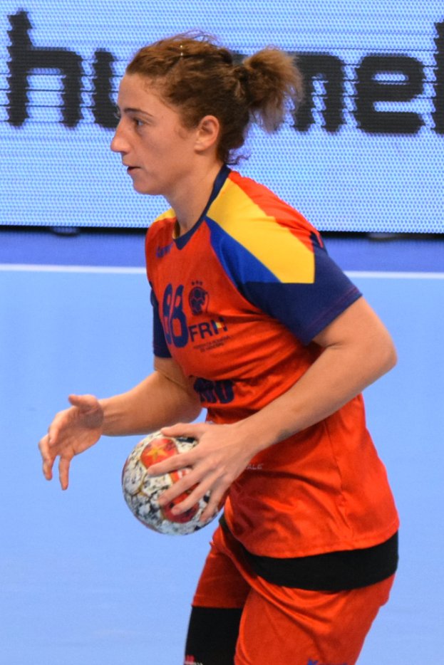 Eine Frau in einer orange-blauen Uniform hält einen Fußball, mit einem Bildschirm im Hintergrund, auf dem Text angezeigt wird.