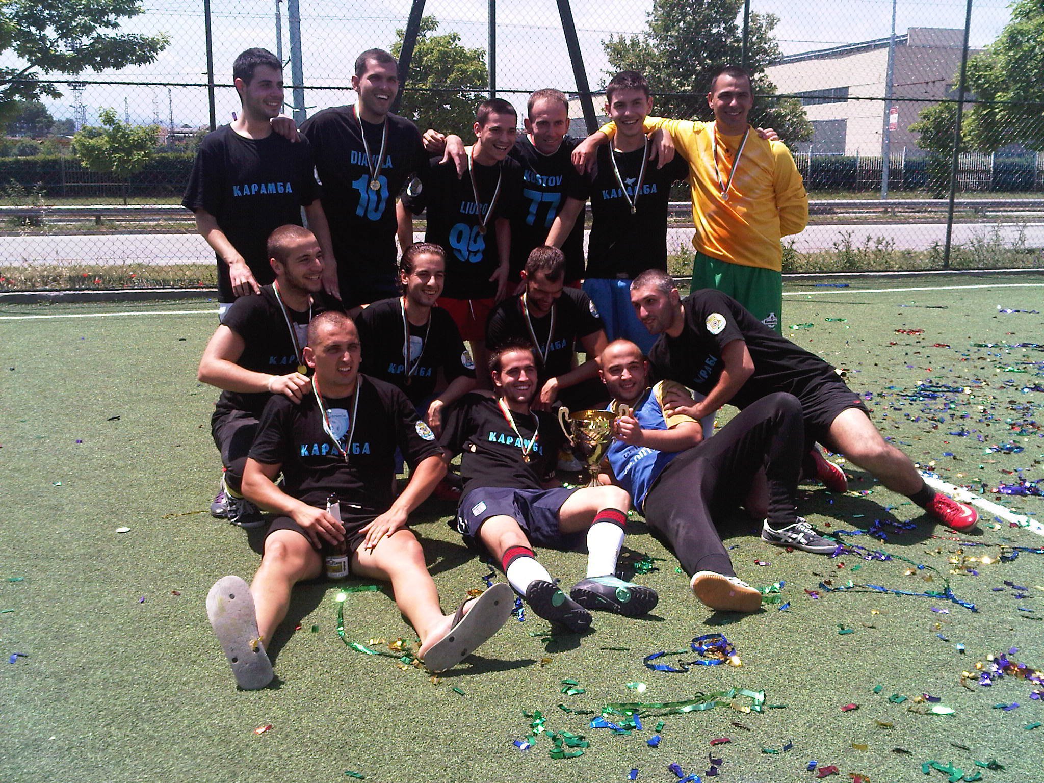 Eine Gruppe von Männern posiert auf einem Rasenfussballfeld für ein Foto, einige tragen Medaillen und einer hält eine Trophäe, mit Maschendrahtzaun, Bäumen, Pflanzen, Gebäuden und einem klaren blauen Himmel im Hintergrund.