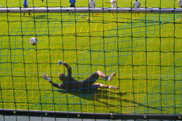 Ein Fußballspieler macht eine Parade vor einem Tor, mit einem Netz im Vordergrund und einigen Menschen im Hintergrund, sowie mehreren Bällen auf dem Boden verstreut.