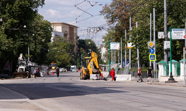 Eine Stadtstraße mit einer Baustelle in der Mitte, mit Fahrzeugen, Fußgängern, einem Radfahrer, Verkehrsleitkegeln, Pfählen, Hinweisschildern, Strommasten mit Drähten, Bäumen, Gebäuden mit Fenstern und einem bewölkten Himmel im Hintergrund.