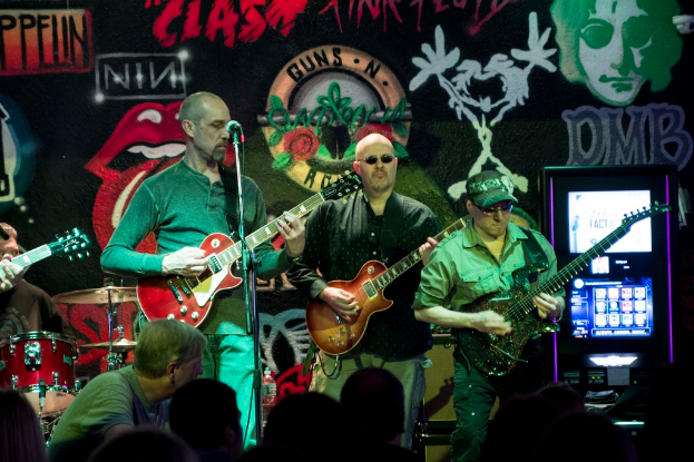 Drei Männer spielen Gitarren auf einer Bühne bei einem Konzert, mit einer Menge unten und einer gemalten Wand im Hintergrund.