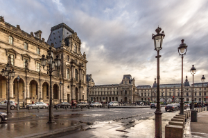 Außenansicht des Louvre-Museums in Paris mit seiner ikonischen Architektur, Straßenlaternen, Lichtern, fahrenden Fahrzeugen, Fußgängern auf dem Gehweg und einem bewölkten Himmel.