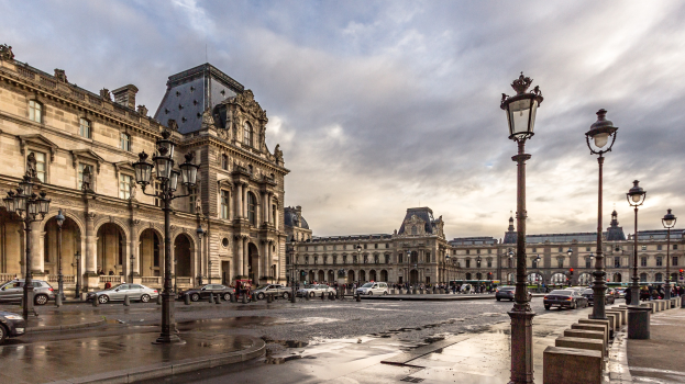 Außenansicht des Louvre-Museums in Paris mit seiner ikonischen Architektur, Straßenlaternen, Lichtern, fahrenden Fahrzeugen, Fußgängern auf dem Gehweg und einem bewölkten Himmel.