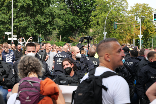 Eine große Gruppe von Menschen, einige mit Masken und Rucksäcken oder Kameras, steht vor einer Menge von Polizeibeamten, mit Laternenmasten, Verkehrsampeln, Verkehrszeichen, Bäumen und einem klaren blauen Himmel im Hintergrund.