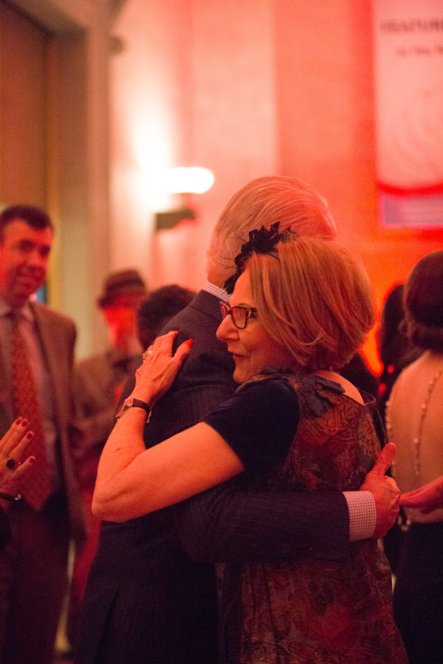 Zwei Menschen umarmen sich auf einer Party, umgeben von anderen und einer beleuchteten Wand im Hintergrund.