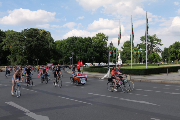 Eine Gruppe von Menschen, die auf Fahrrädern eine von Bäumen gesäumte Straße mit Laternenmasten und Flaggenmasten entlangfahren, während sie an der Pariser Radtour 2016 teilnehmen.
