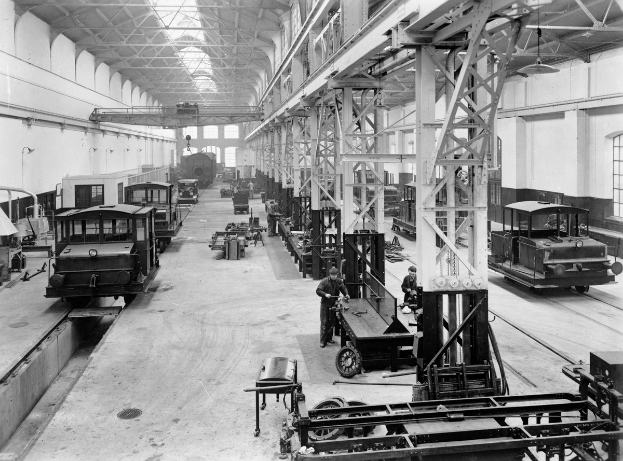 Schwarzes und weißes Foto einer Fabrikhalle mit mehrerenbahnen in Reparatur, Arbeitern, die sich darum kümmern, Metallpfosten, die die Konstruktion stützen, Deckenlampen und Fenstern an den Wänden.