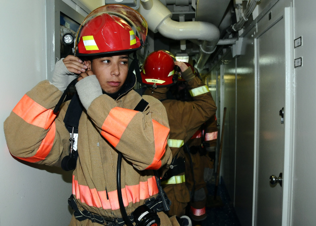 Feuerwehrleute in Uniform stehend in einem Raum während einer Übung, mit Rohren und Equipment im Hintergrund.