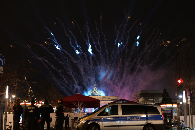 Ein Polizeiwagen steht vor einer Menschenmenge, umgeben von Fahrrädern, Zelten, Laternenmasten, Schildern, Verkehrszeichen, Bäumen, Gebäuden und einer Statue im Hintergrund, unter einem von Feuerwerk erhellten Himmel.