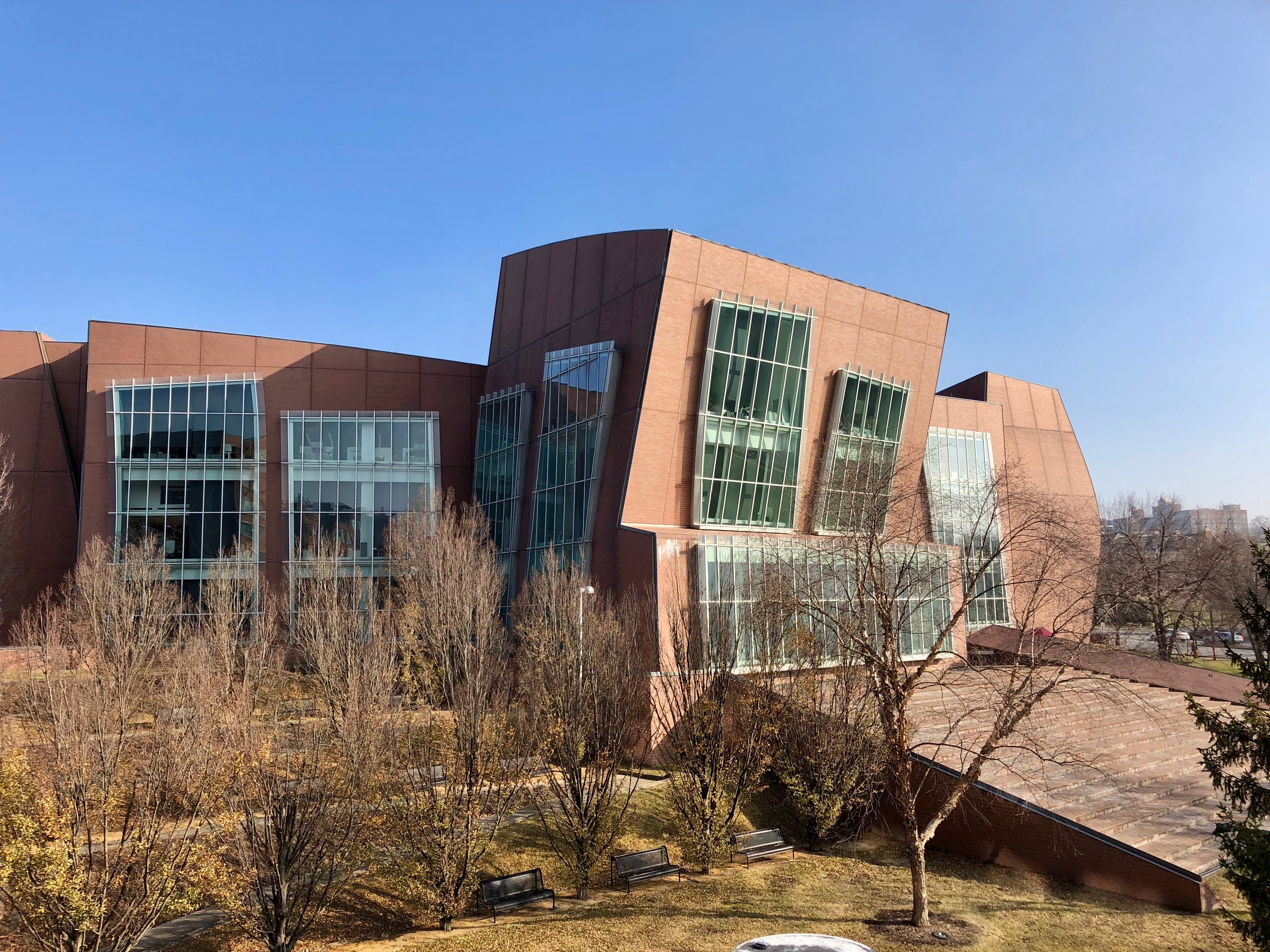 Großes Gebäude mit vielen Fenstern, umgeben von Bäumen, Bänken, Gras und einem Laternenmast, vor einem klaren blauen Himmel mit anderen Gebäuden im Hintergrund.