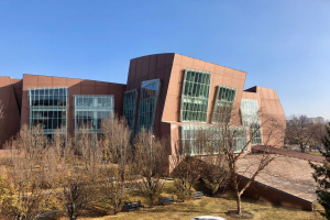 Großes Gebäude mit vielen Fenstern, umgeben von Bäumen, Bänken, Gras und einem Laternenmast, vor einem klaren blauen Himmel mit anderen Gebäuden im Hintergrund.