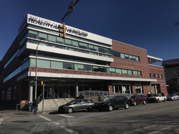 Großes Gebäude mit der Aufschrift "New York City Health Care Homeless Center" und parkenden Autos davor, umgeben von Straßeninfrastruktur und Passanten unter einem klaren blauen Himmel.