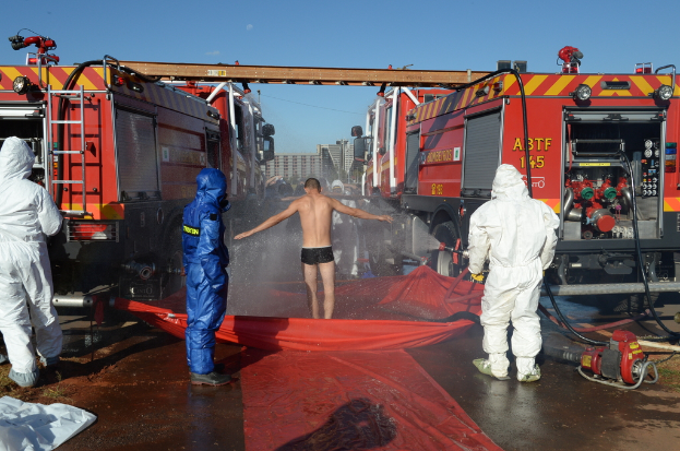 Drei Personen in Warnwesten stehen im Vordergrund, eine Person steht auf einer Abdeckung mit Wasser davor. Einige andere sind in der Nähe, und Feuerwehrautos mit ausgefahrenen Metallstangenleitern sind daneben positioniert. Gebäude sind im Hintergrund zu sehen, und es gibt eine Abdeckung mit einigen Gegenständen auf der Oberfläche.