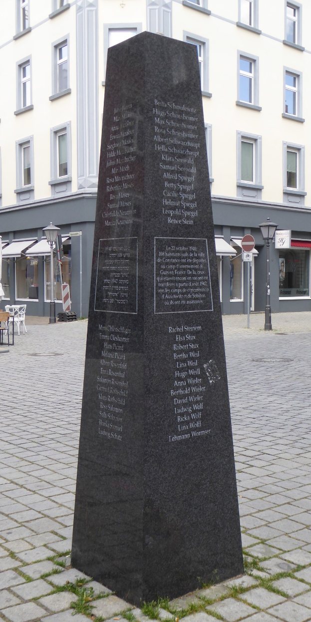 Ein hohes Denkmal mit eingravierter Inschrift steht auf einem Platz in der Stadtmitte, umgeben von Gebäuden, Straßenmöbeln und Menschen unter einem sichtbaren Himmel.