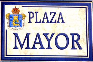 Rechteckige weiße Plakette mit schwarzem Text "Plaza Mayor" in fetter Schrift, blauem Rahmen und einem zentralen gekrönten Wappen-Logo an der Seite eines Gebäudes.
